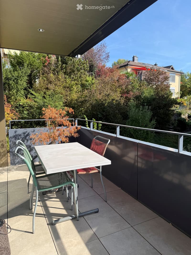 Balcony with a white table and two chairs, surrounded by greenery and a modern railing.