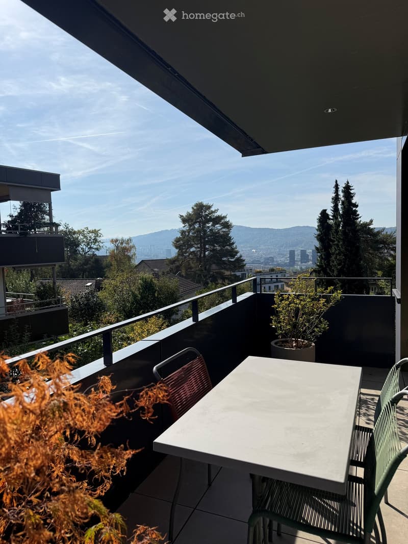 Balcony with a table and chairs, overlooking a cityscape with trees and mountains in the background.