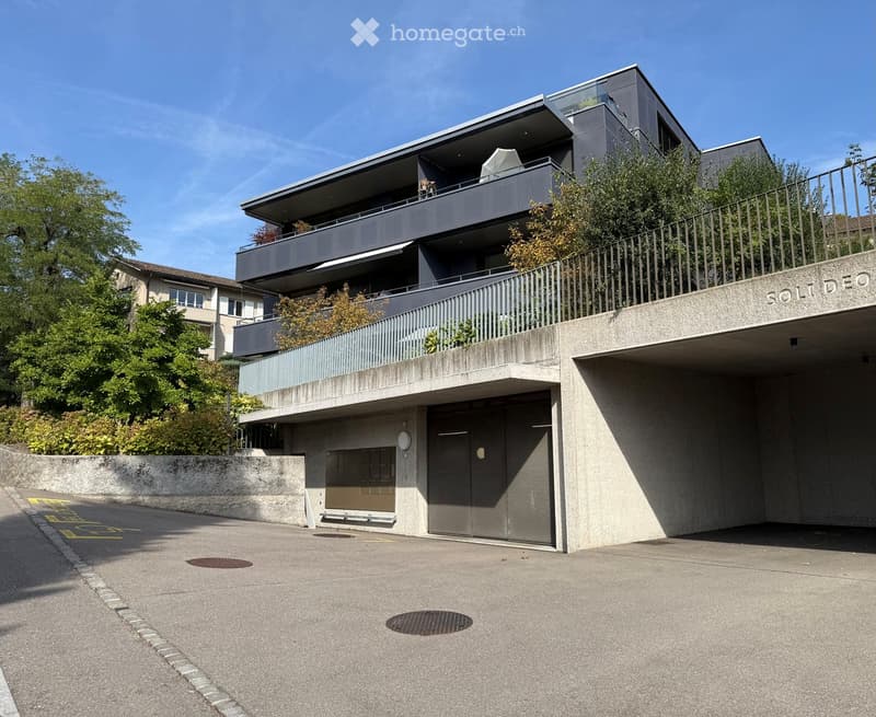 Modern multi-story building with a garage entrance, balconies, and greenery on the upper levels.