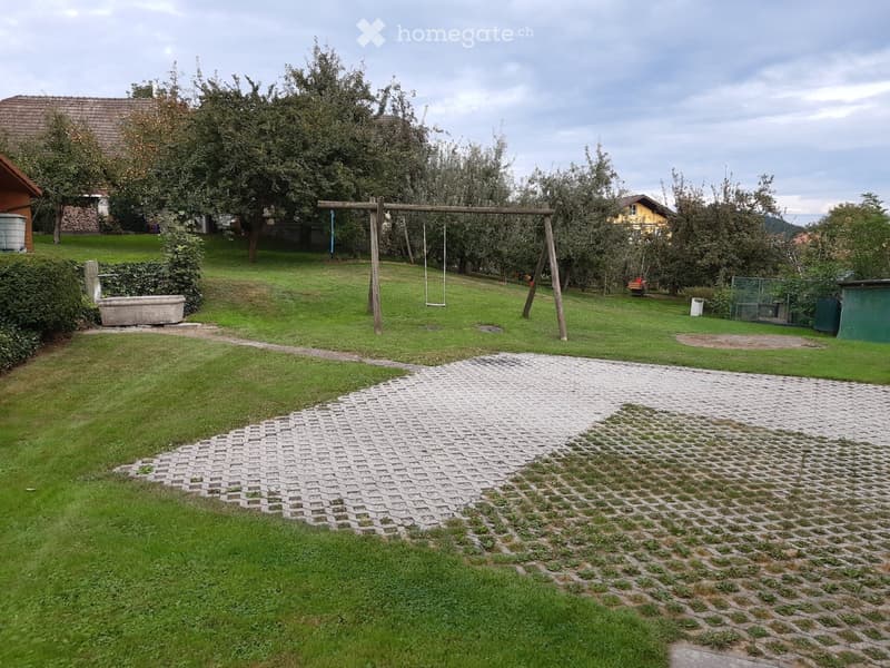 A well-maintained garden with a paved pathway, grassy areas, trees, a swing set, and a small shed.