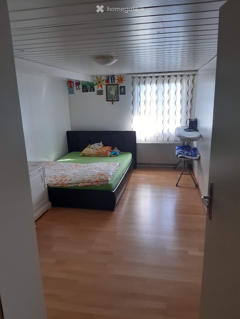 A small bedroom with a single bed, pillows, a blanket, a window with curtains, and a small table with items on it.