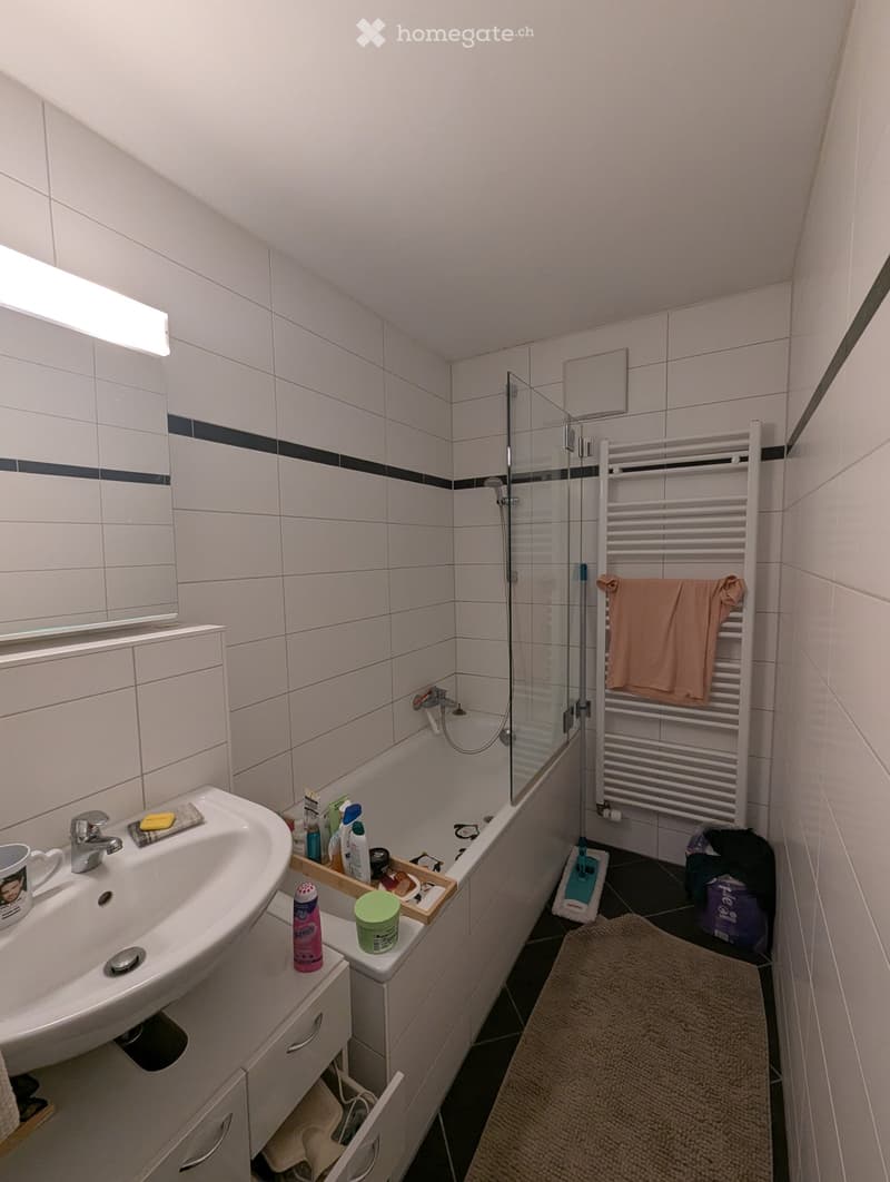 Modern bathroom with white tiles, a sink with a mirror, a shower area, a towel rack with a towel, and a bathroom tray with various items on the counter.