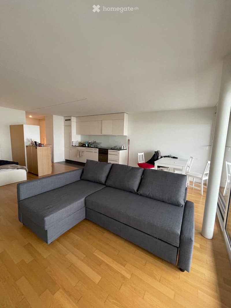 A modern, open-concept living space with a grey sectional sofa, a small dining table with chairs, and a compact kitchen area featuring light-colored cabinets, an oven, and a refrigerator.