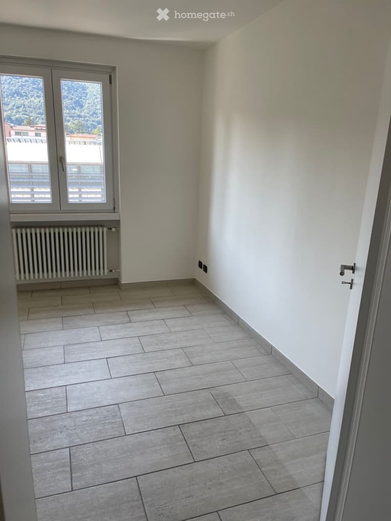 Empty room with tiled floor, white walls, large window with blinds, radiator, and door.