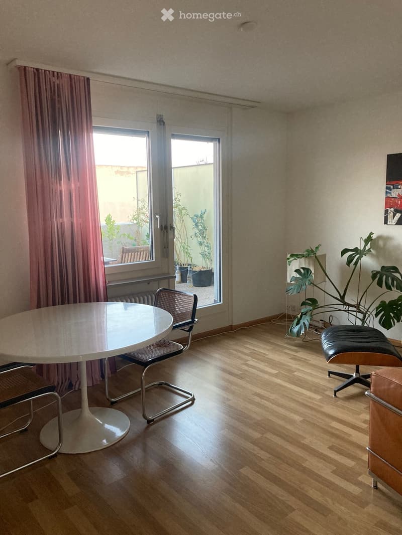 Dining area with a round white table, chairs, and a leather sofa, with large windows and plants visible outside.