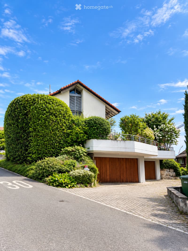 Modern two-story house with a flat roof, wooden garage doors, and a well-maintained garden with trimmed hedges and shrubs.