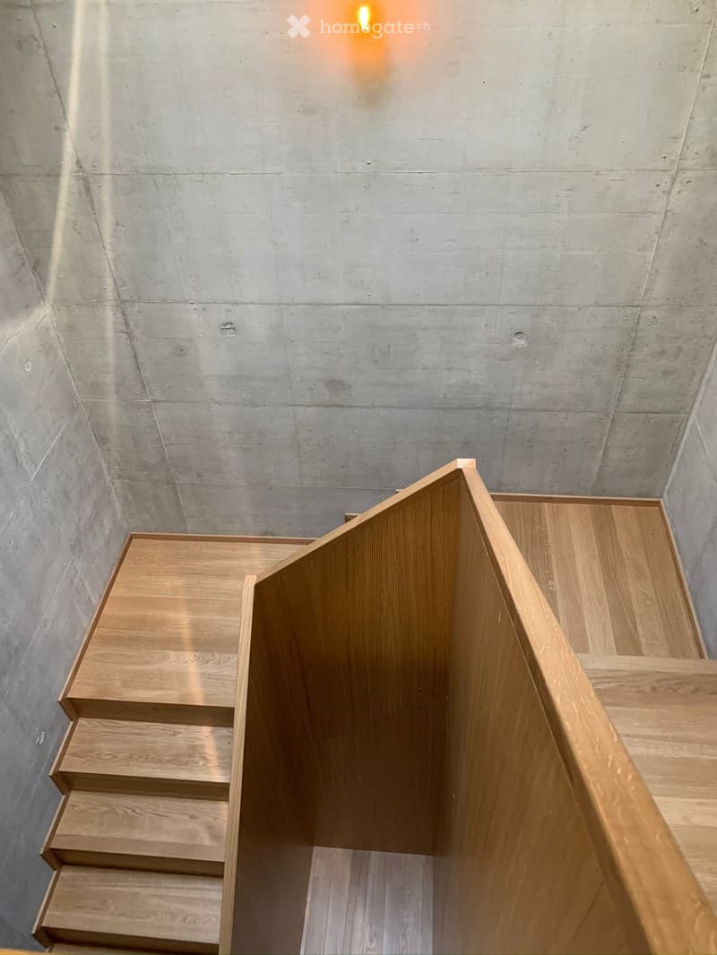 Modern staircase with wooden steps and railings, concrete walls, and a warm ambient light fixture.