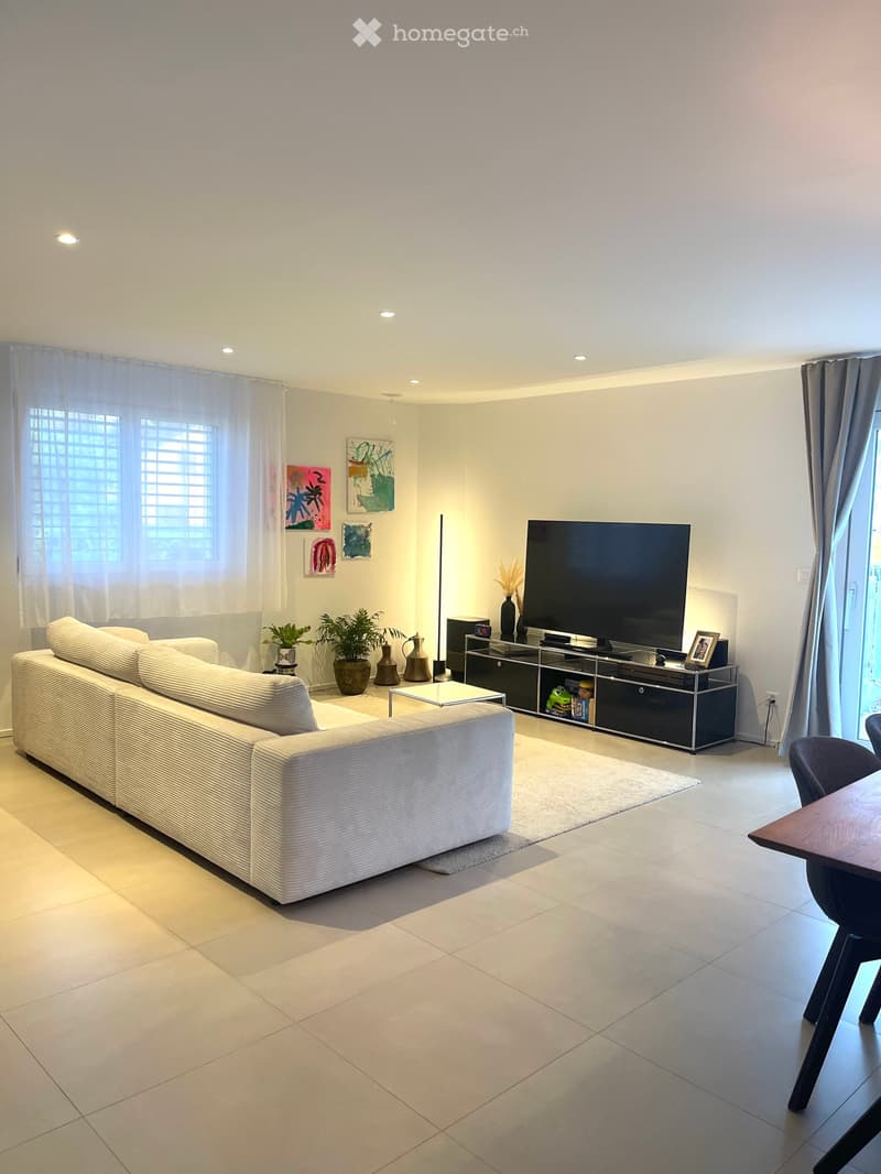 Modern living room with a white sectional sofa, coffee table, large flat-screen TV, and decorative vases. The space features large windows with sheer curtains and a tiled floor.