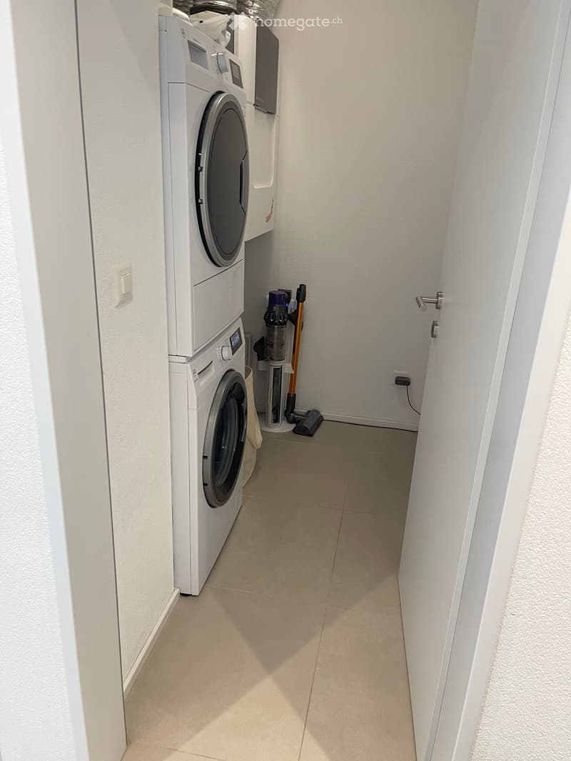 A narrow hallway with a private washing machine and dryer unit on the left side, a vacuum cleaner and its accessories on the floor, and a door on the right side.