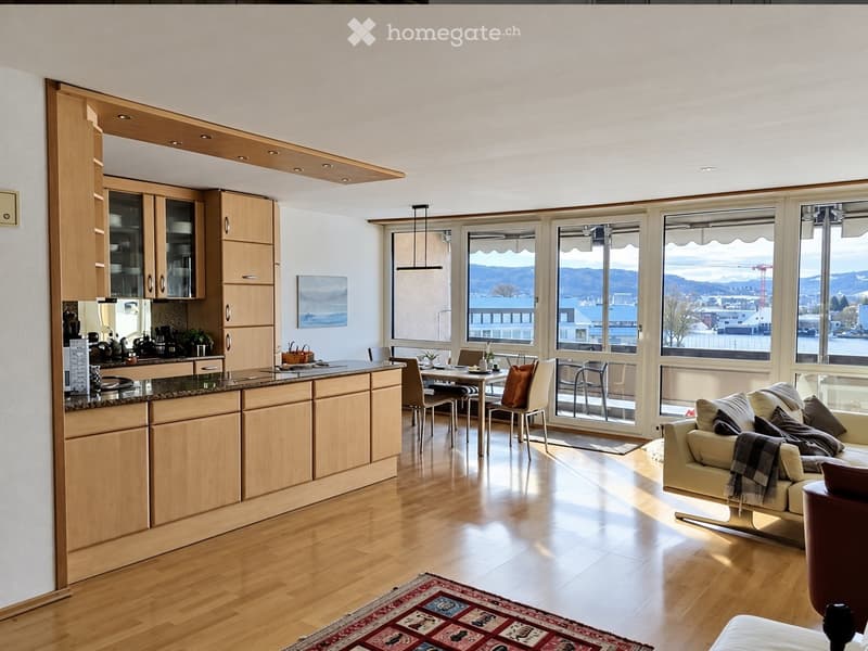 Bright, open-plan living and dining area with wooden flooring, large windows offering a scenic view, a cream-colored sofa, a dining table with chairs, and a modern kitchen with wooden cabinets and a granite countertop.