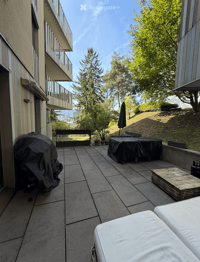 Balcony with tiled floor, hot tub, covered table, umbrella, and bench, overlooking a landscaped area with trees and a city view.