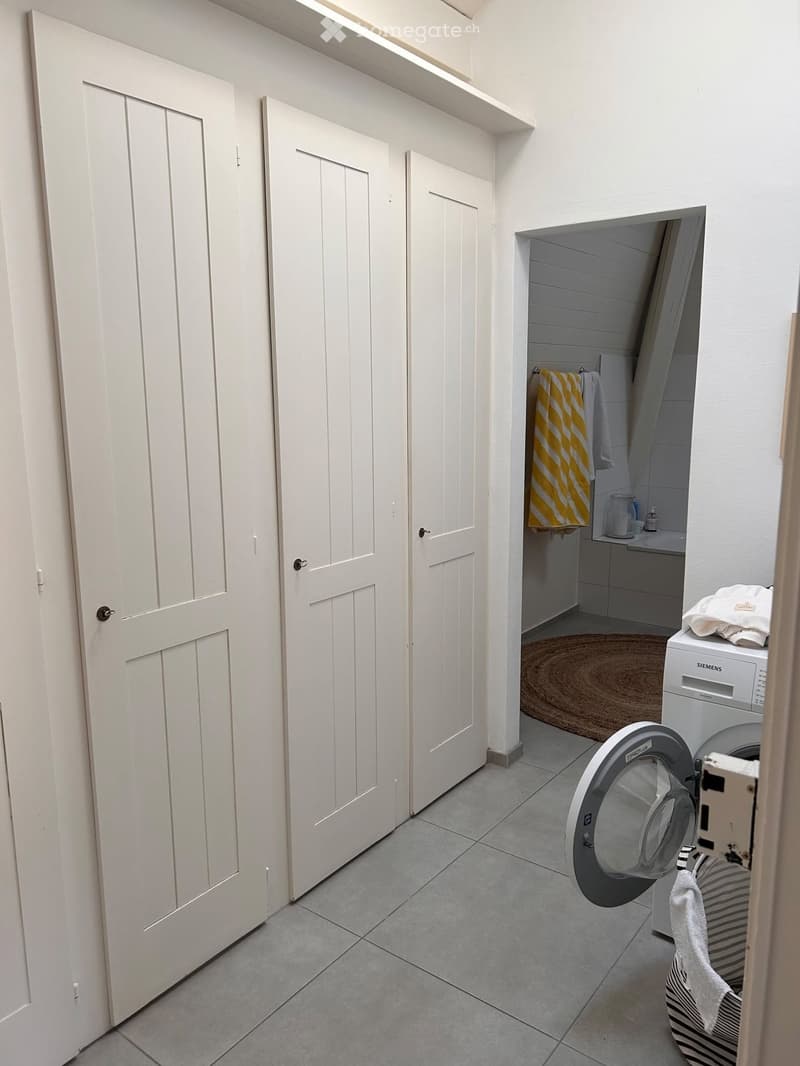 A small bathroom with a washing machine, a sink, and a towel hanging on the wall. The room has white doors and tiled flooring.