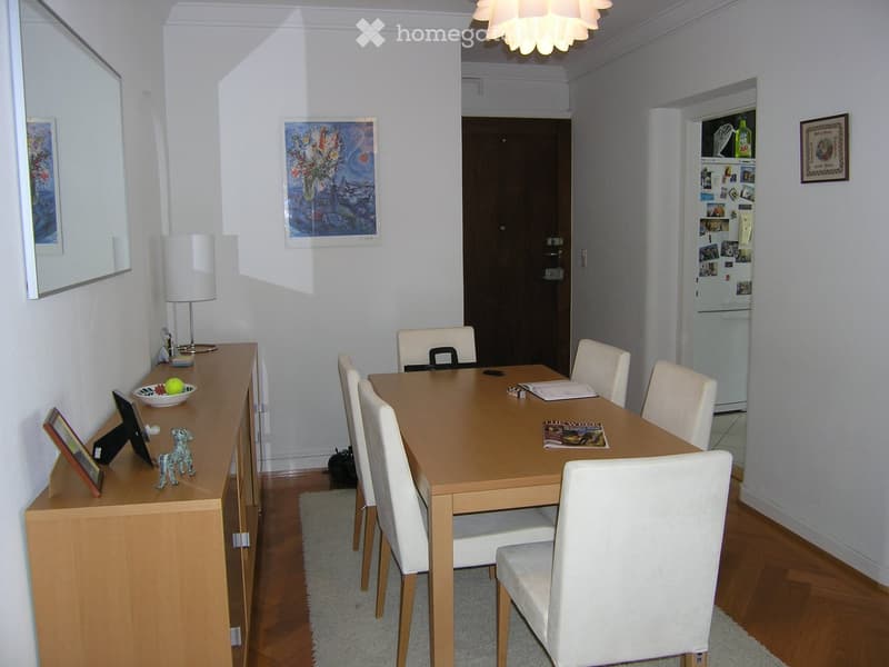 Dining room with a wooden dining table, six white chairs, a sideboard with a lamp, a mirror, and a refrigerator in the background.