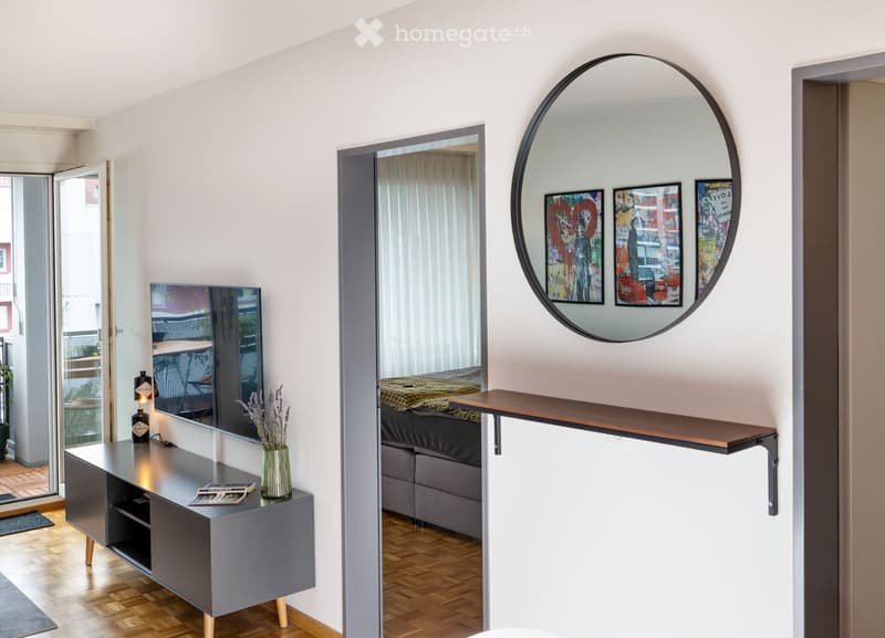 Modern living room with a grey console table, a round mirror, a television, a sofa, and a vase with lavender flowers. The room features wooden flooring and large windows with white curtains.