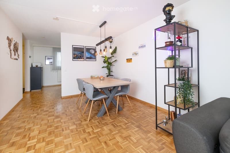 Modern dining area with a wooden dining table, four grey chairs, a tall black shelving unit with plants and decorative items, and framed pictures on the wall. The room features wooden herringbone flooring and pendant lighting.