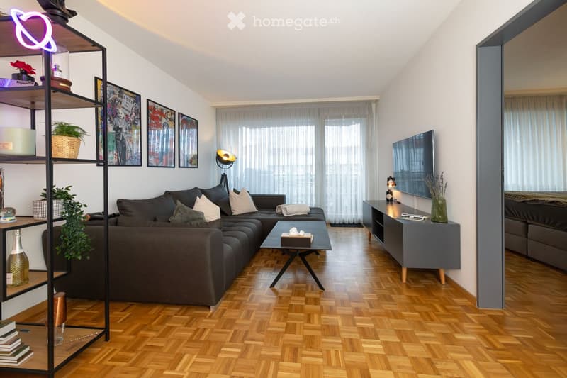Modern living room with wooden flooring, large sectional sofa, coffee table, side table, wall-mounted TV, decorative shelf, and artwork.