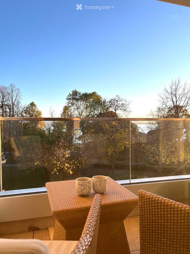 Balcony with a wicker table, two chairs, and two cups, overlooking a tree-lined area with a clear blue sky.