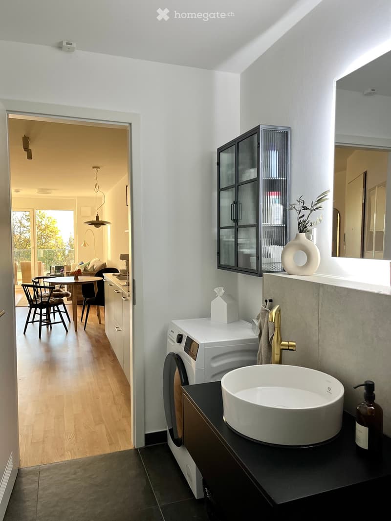 Modern bathroom with a white sink, gold faucet, washing machine, and a large mirror. A black cabinet holds a vase and a tissue box. The adjacent room features a dining area with chairs and a table.
