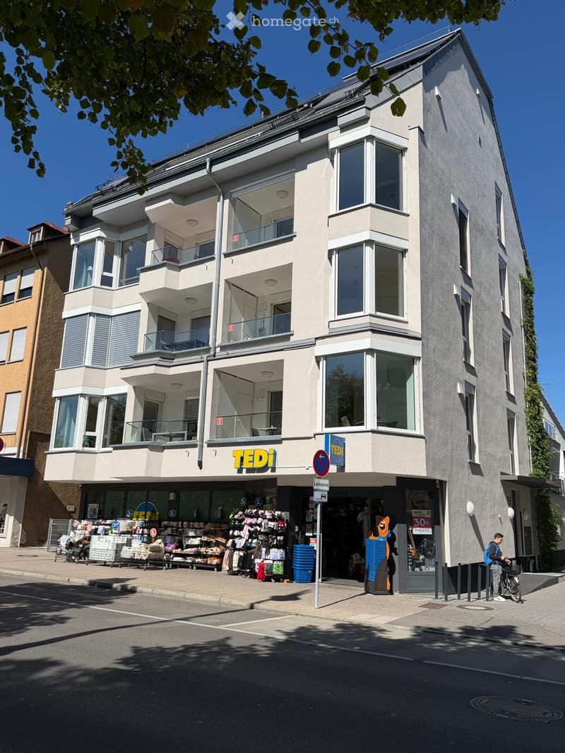 Modern multi-story building with large windows and balconies, featuring a storefront with signage reading "TEDi" and various displayed items outside.