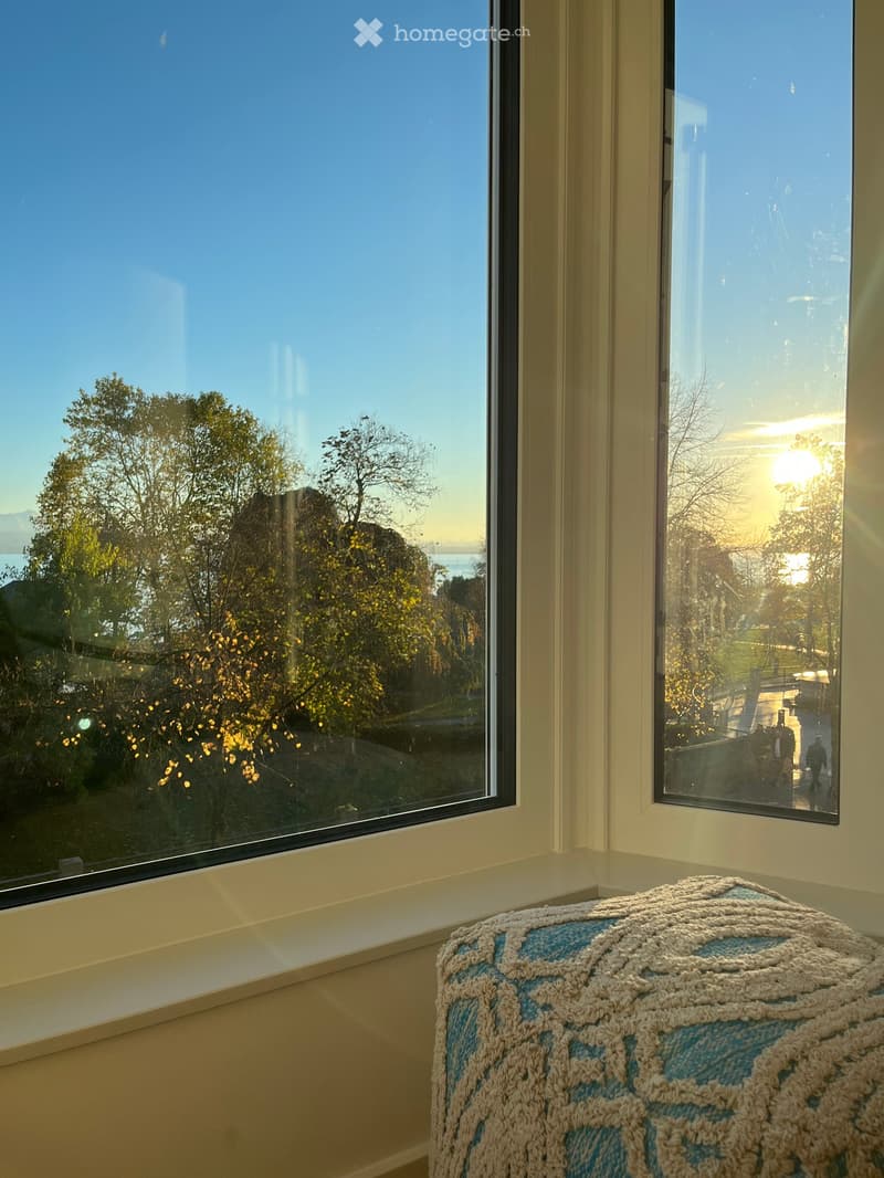 A cozy room with large windows offering a scenic view of trees and a body of water. A blue and white crocheted stool sits near the windows.