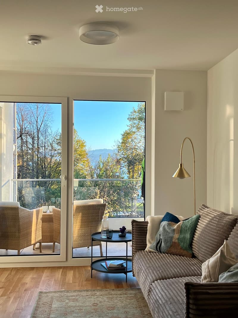 Modern living room with large windows offering a scenic view, featuring a sofa, coffee table, floor lamp, and a small table with decorative items.