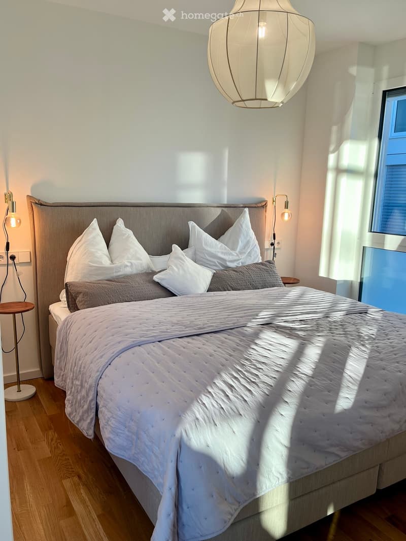Modern bedroom with a large bed, white bedding, grey headboard, and two side lamps. Natural light streams in from a window on the right.
