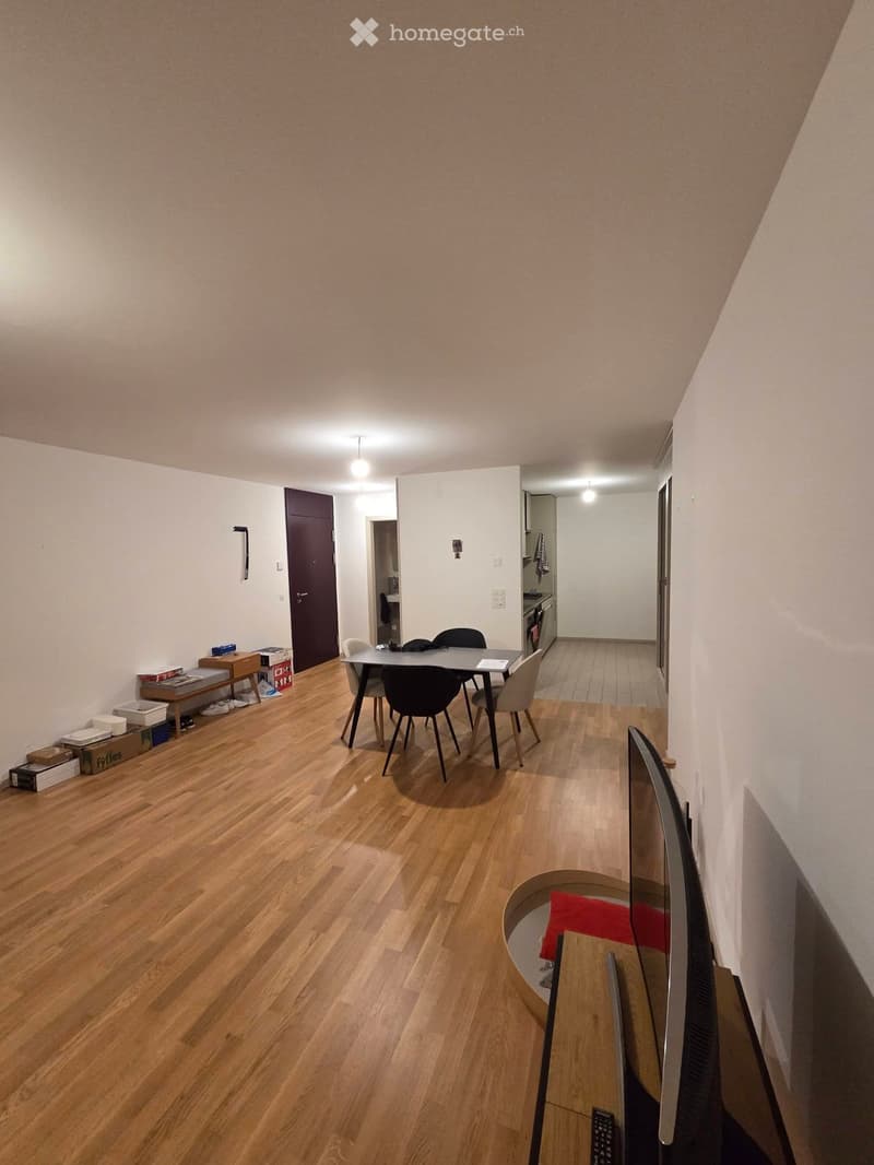 A furnished room with wooden flooring, a small dining area with a table and chairs, a TV on a stand, and a small bench with boxes and items.