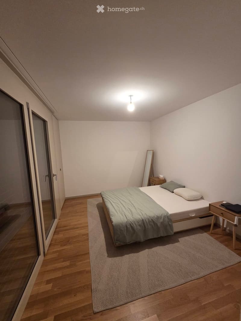 Modern bedroom with a single bed, nightstand, mirror, and laptop on the nightstand. The room features wooden flooring and a large window with a glass door.