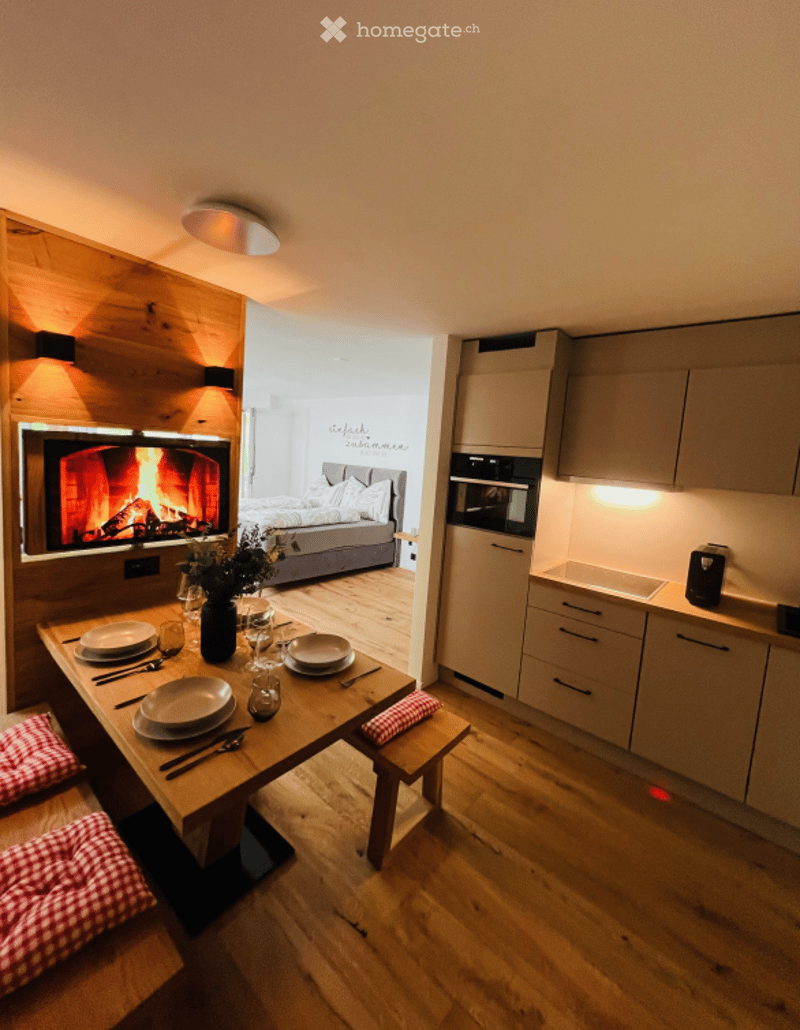 A modern, open-plan living and dining area with a central wooden dining table, fireplace, sofa, and a small kitchen area with cabinets and an oven.