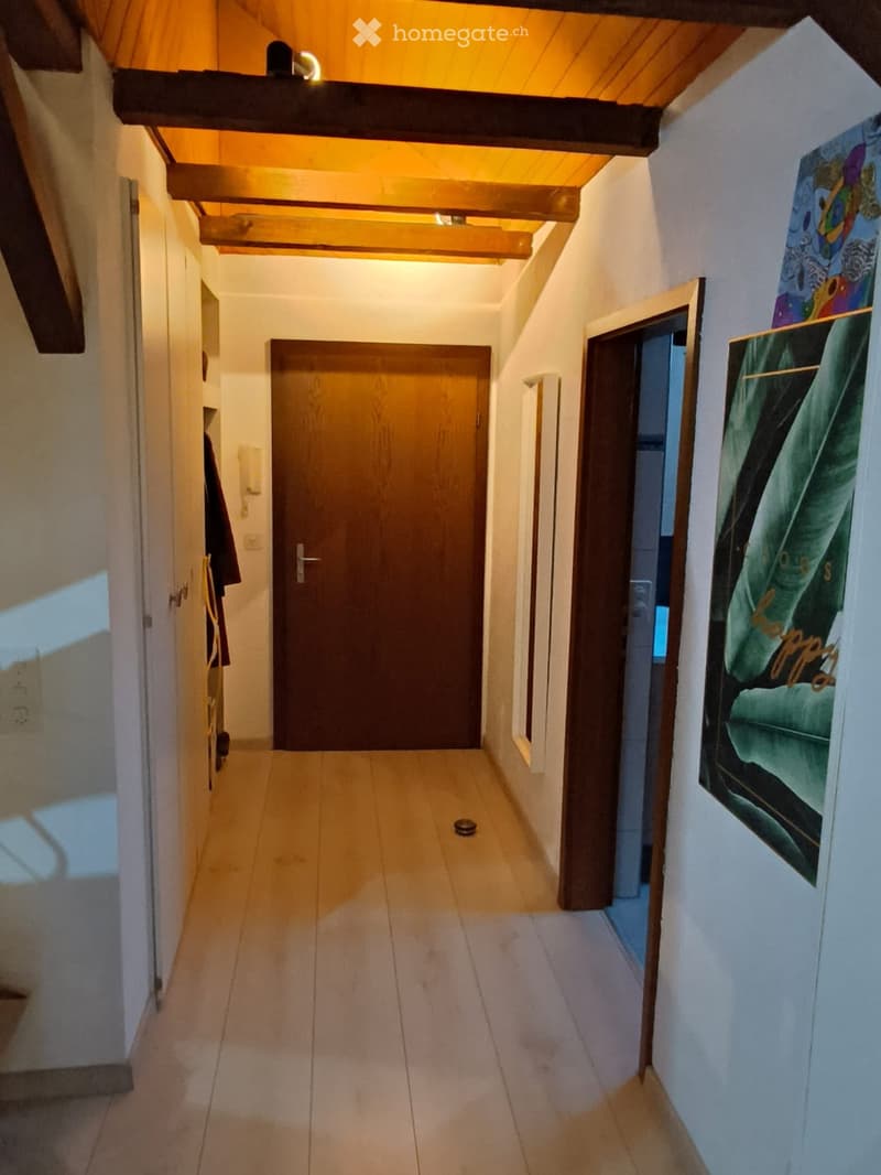 A narrow hallway with wooden beams on the ceiling, tiled floor, and several doors leading to different rooms. There are framed pictures on the right wall and a coat hanging on the left side.