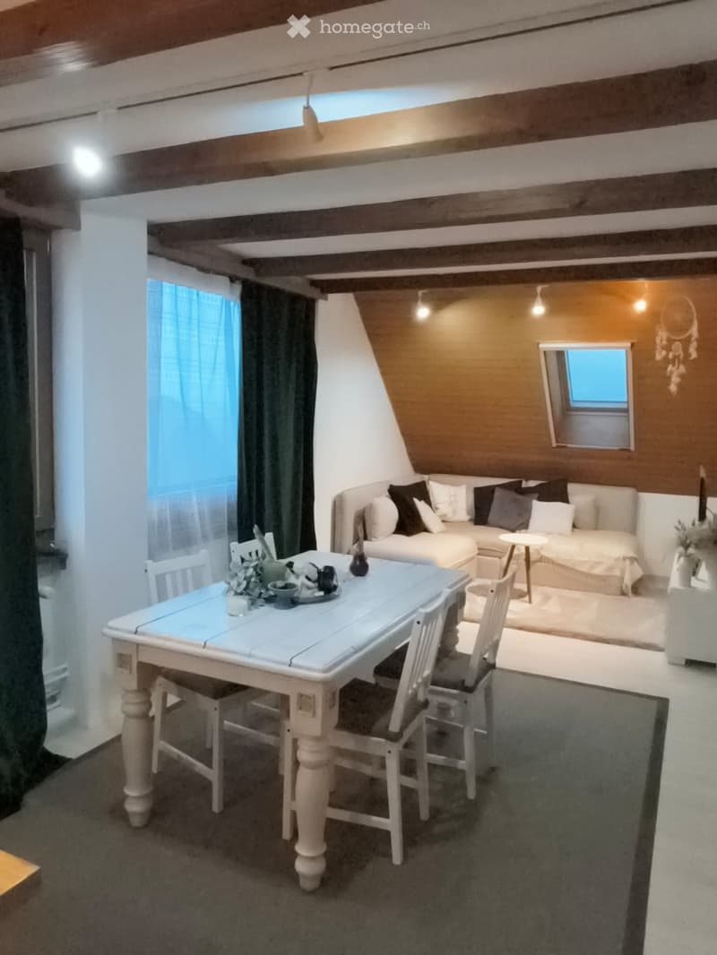 Cozy attic room with exposed wooden beams, a sofa, a dining table with chairs, and a small mirror on the wall.