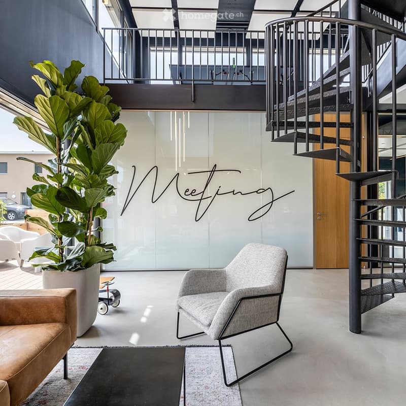 Modern, open-plan meeting space with a large potted plant, a grey armchair, a brown sofa, a small table, and a spiral staircase leading to an upper level.
