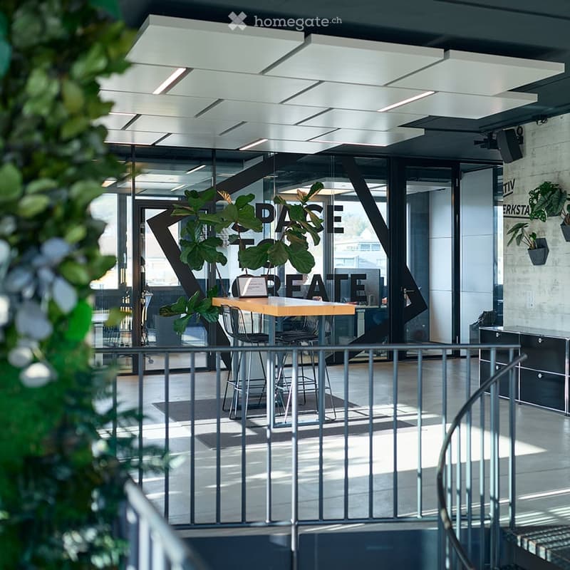 Modern, well-lit space with a wooden table, chairs, and a large plant, featuring a sleek, contemporary design with glass walls and a metal railing.