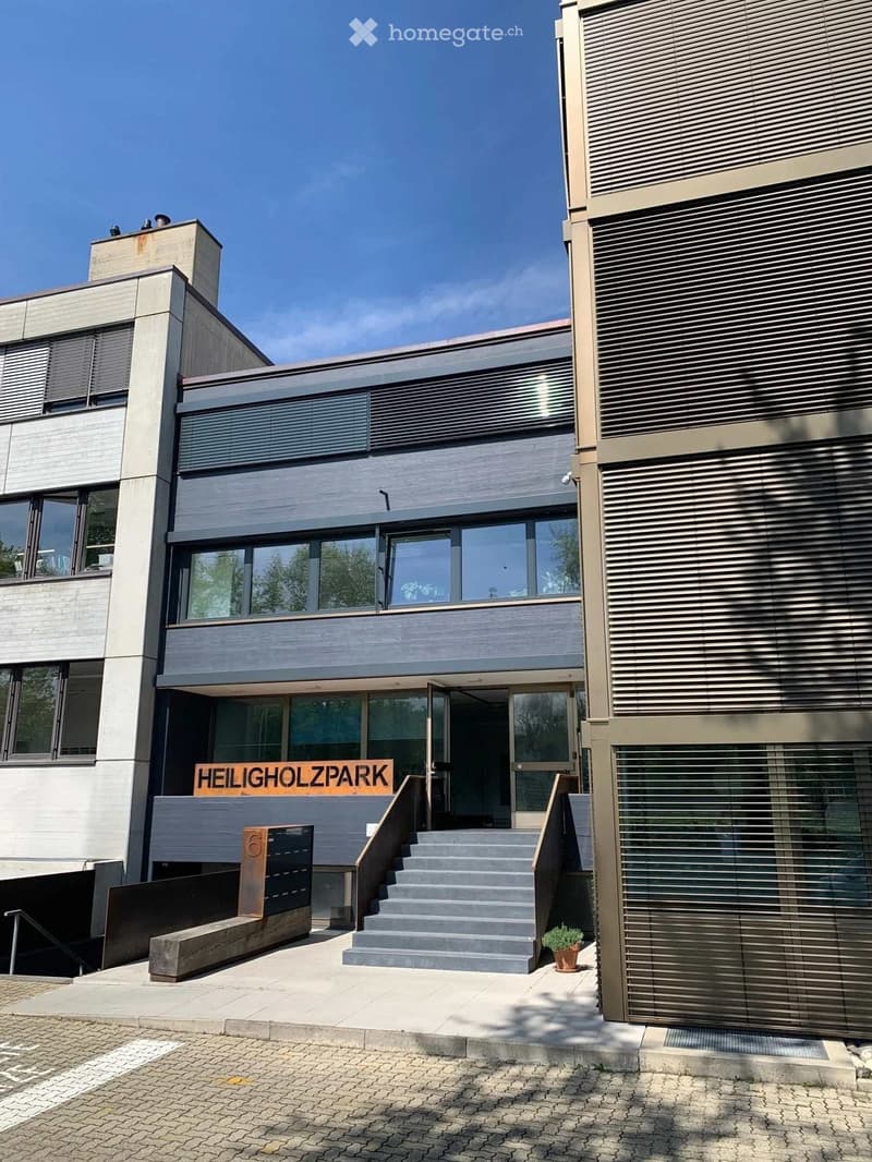 Modern two-story building with large windows, grey facade, and a staircase leading to the entrance. The building features a sign that reads "HEILIGHOLZPARK" and a small potted plant near the steps.