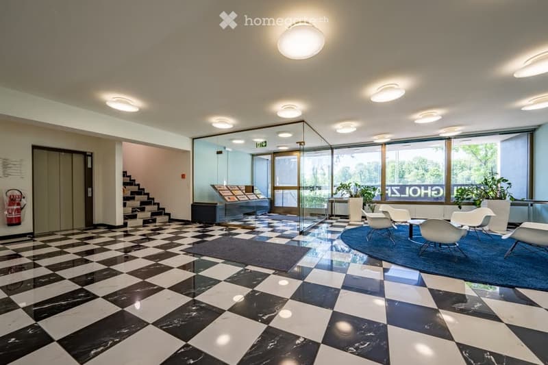 Modern reception area with black and white checkered marble flooring, white chairs around a small table, large windows with a view of greenery, and a staircase leading upwards.