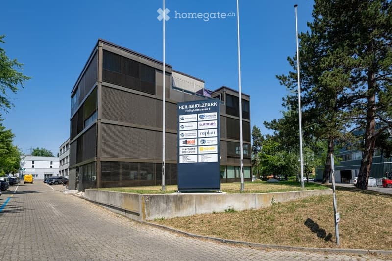 Modern multi-story office building with a signboard displaying company names, surrounded by trees and a paved driveway with parked cars.