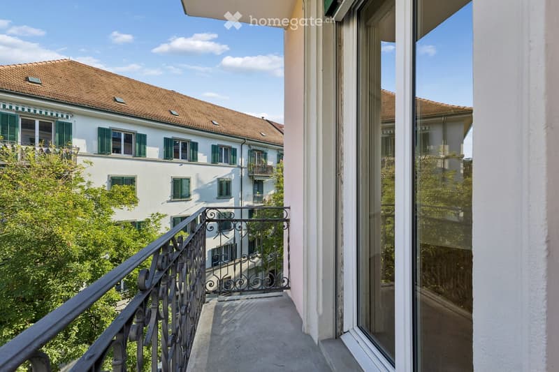 Balcony with metal railing overlooking a tree-lined street and neighboring buildings with green shutters and awnings.