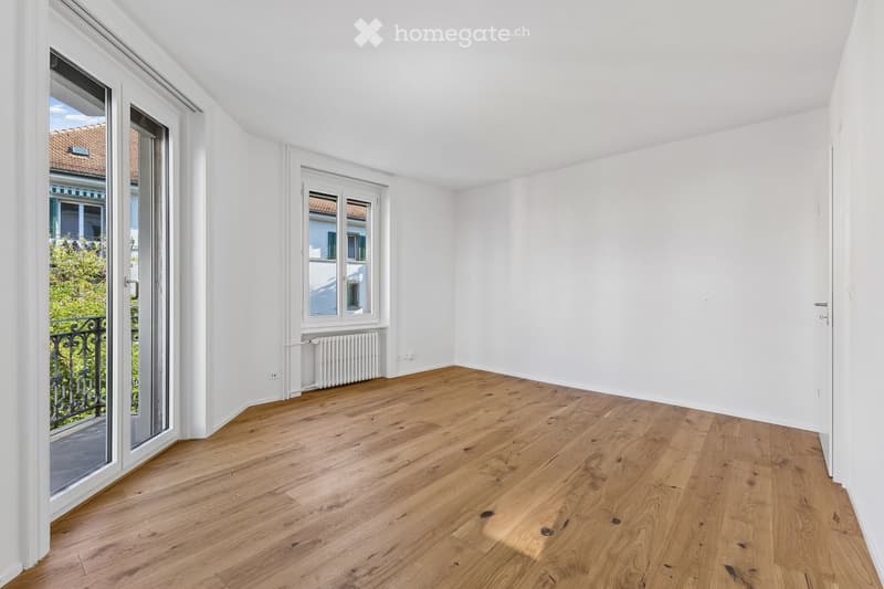 Empty room with light wooden flooring, white walls, large window, and sliding glass door leading to a balcony.