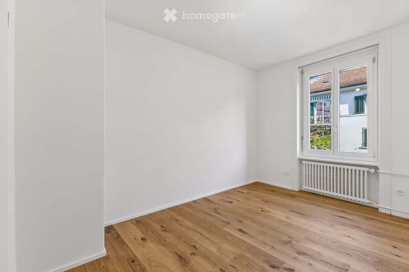 Empty room with white walls and wooden flooring, featuring a large window with a radiator below it.