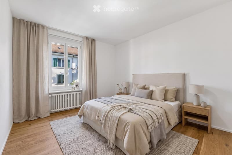 Modern bedroom with a large bed, nightstands, lamps, and a view of a cityscape through large windows.