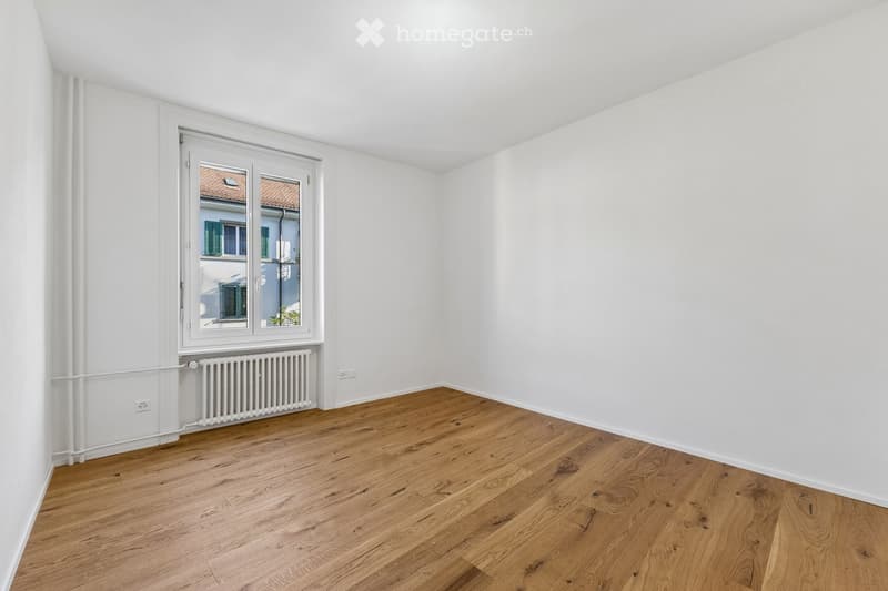 Empty room with light wooden flooring, white walls, and a large window with a radiator below it.
