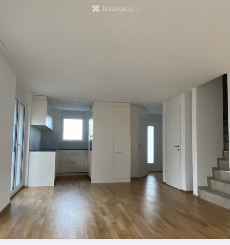 Empty room with wooden flooring, white walls, and a small kitchen area visible in the background. No furniture or equipment present.