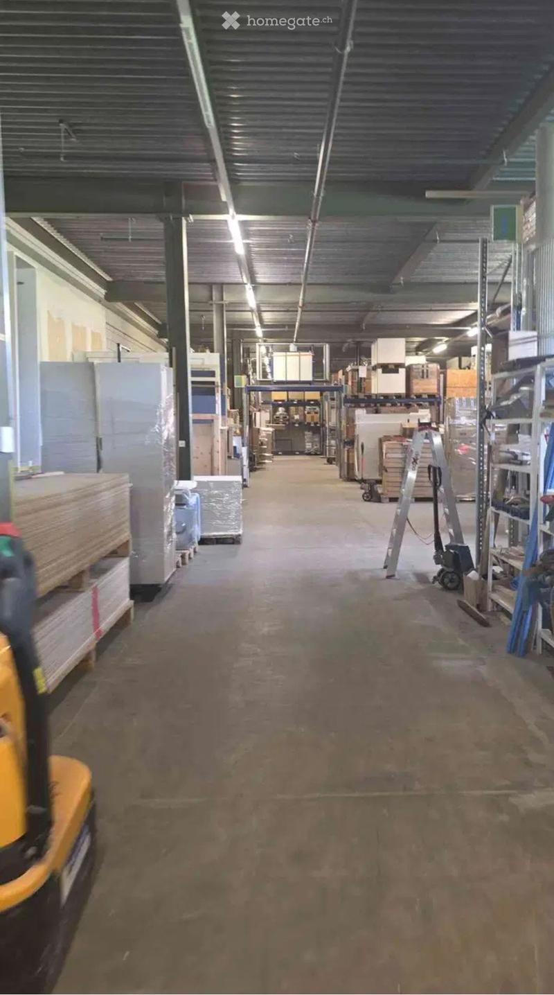 Wide, empty industrial corridor with exposed metal ceiling, fluorescent lighting, and storage racks on both sides.
