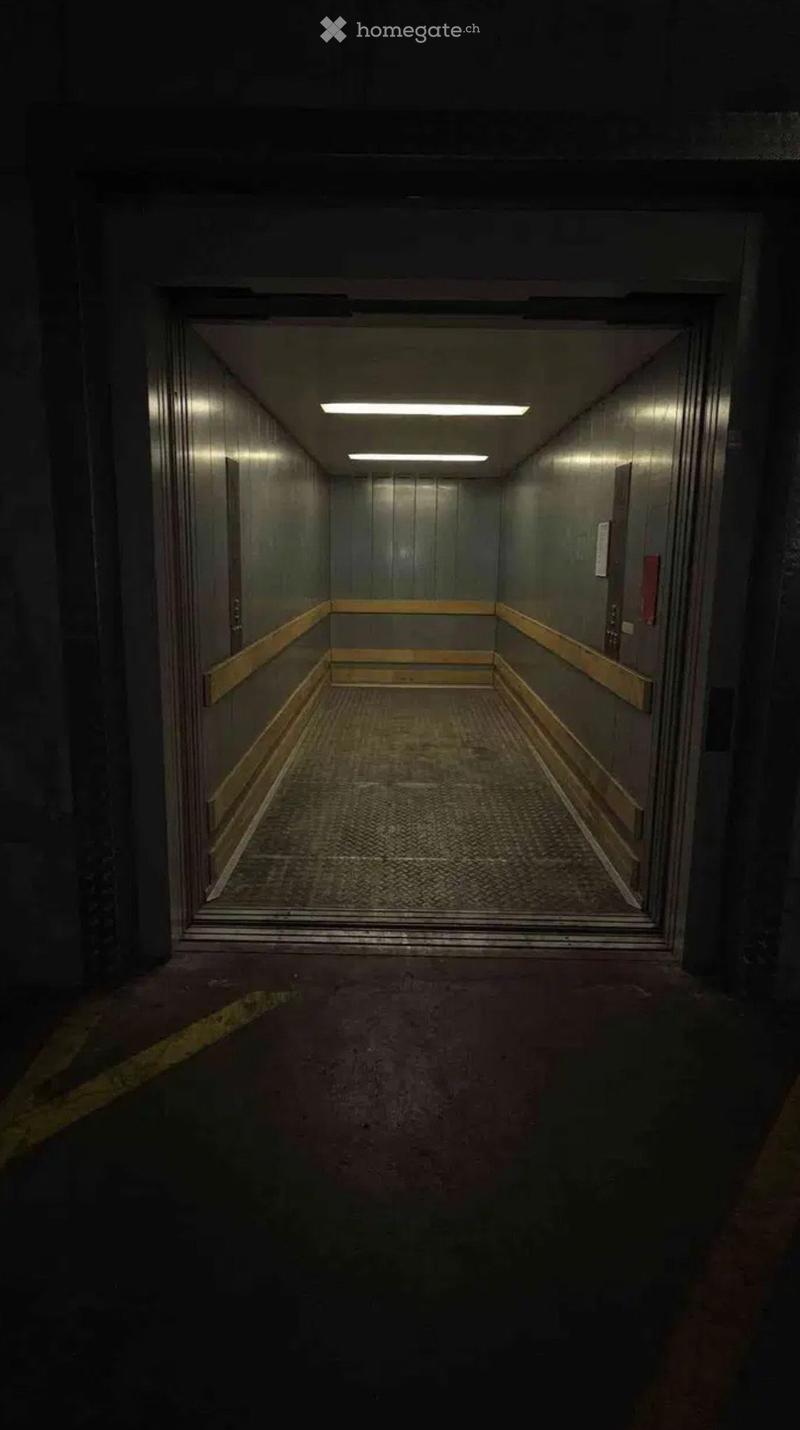 Elevator shaft with metallic walls, a textured floor, and overhead lighting. The elevator doors are open, revealing the interior of the elevator car.