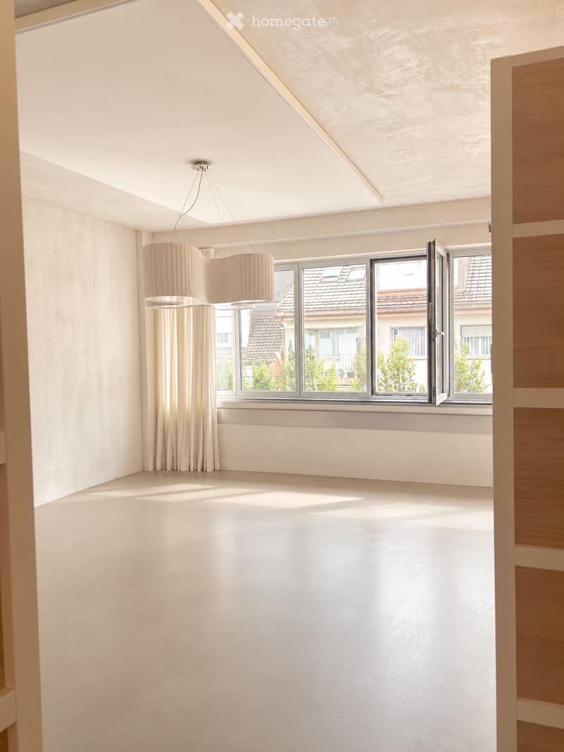 Empty room with white walls, light-colored floor, large window with view of neighboring buildings, and a modern pendant light fixture.