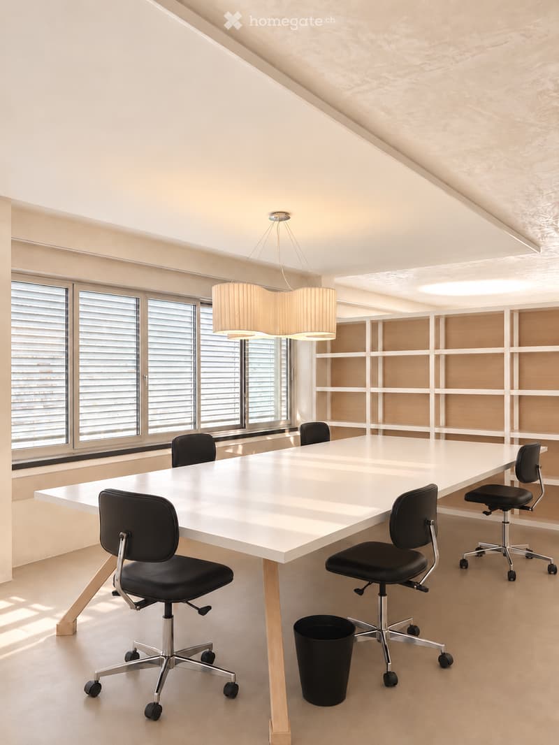 Modern conference room with a long white table, black office chairs, pendant lamps, and built-in shelving units.