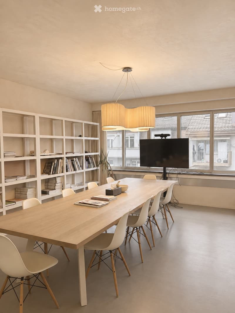 Bright, minimalist room with a long wooden table surrounded by white chairs, a large screen on a stand, and a tall shelving unit filled with books and decorative items.