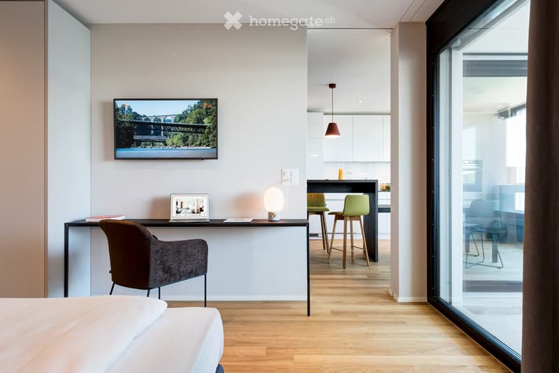 Modern, well-lit room with a bed, desk, chair, laptop, two stools, a TV screen, and a lamp. The room features large windows and a sleek, contemporary design.
