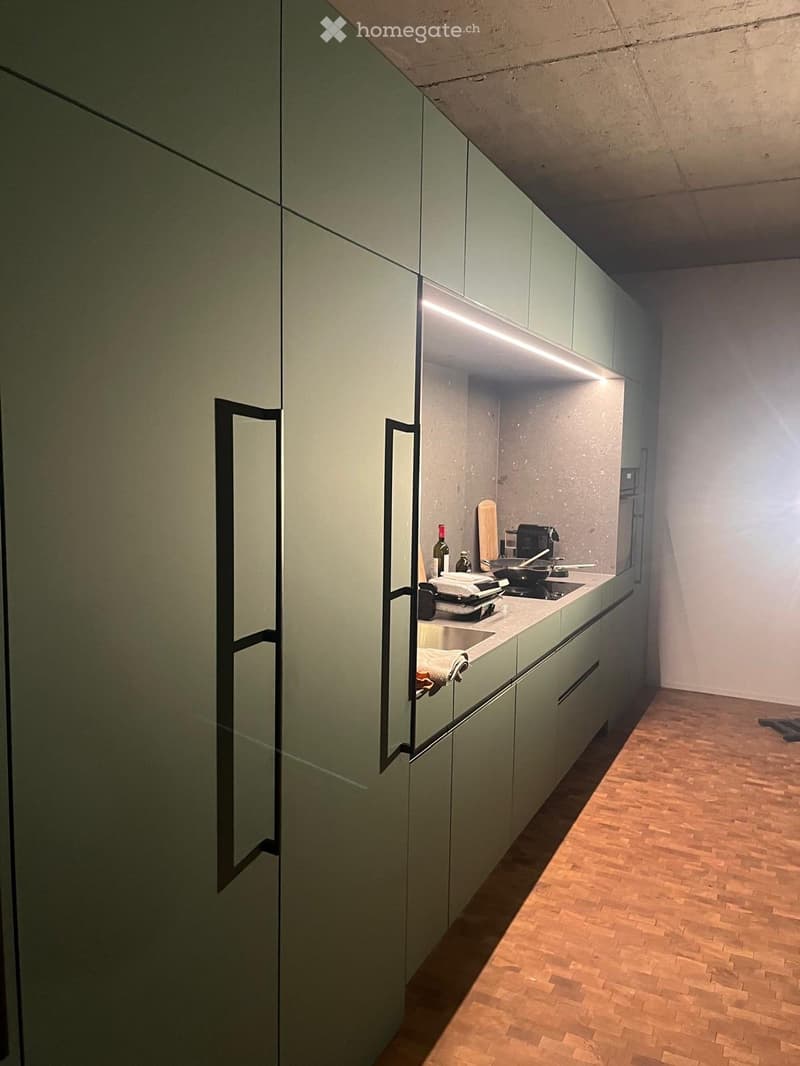 Modern kitchen with light green cabinets, wooden flooring, and built-in appliances including a sink, stove, and refrigerator. A bottle and cutting board are placed on the counter.