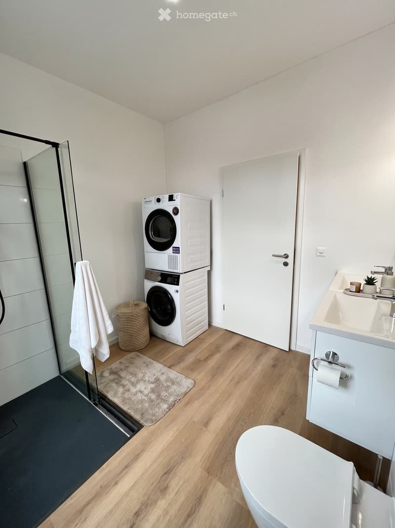 Modern bathroom with white walls, wooden flooring, a sink with a mirror, a toilet, a washing machine and dryer stacked, a glass shower enclosure, a towel, a wicker basket, and a small rug.